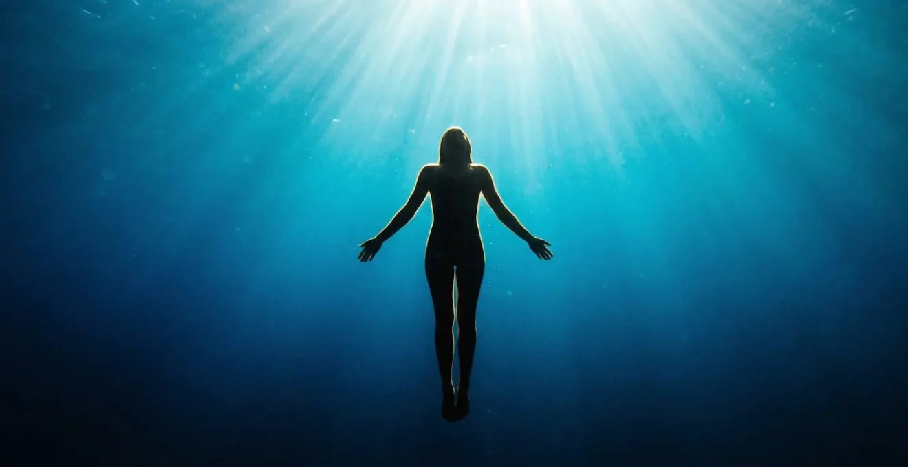 Silhouette humaine en suspension verticale dans l'eau bleue profonde, éclairée par des rayons de soleil naturels depuis la surface