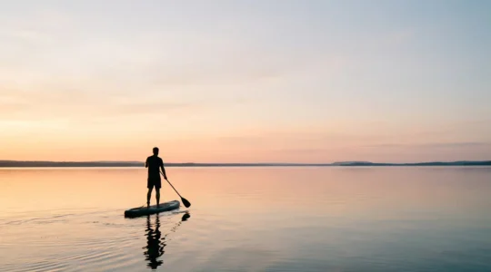 Pratiquant de stand-up paddle sur une eau calme au lever du soleil, vue grand angle avec espace négatif et horizon dégagé